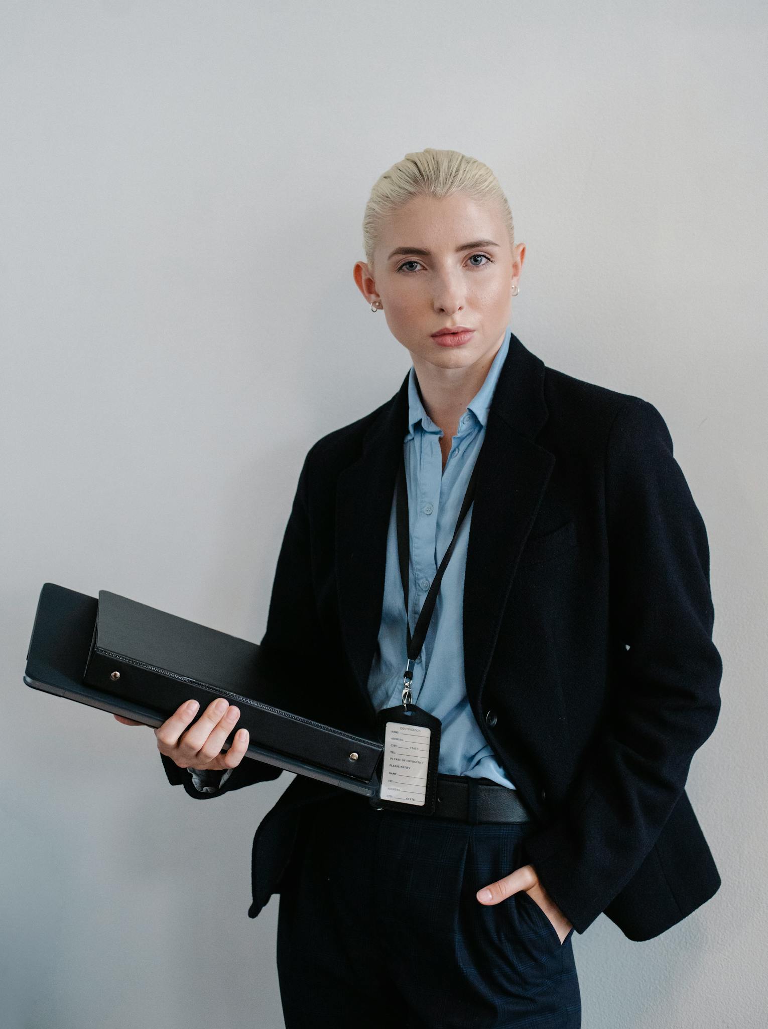 Positive independent businesswoman in black formal clothes standing with hand in pocket and folders in hand against white wall while looking at camera with confidence